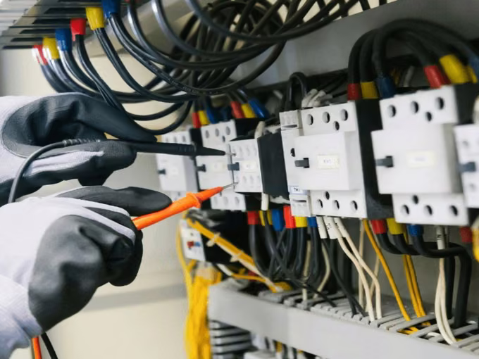 Electrician in gloves testing contactors in a wired control panel with a multimeter probe