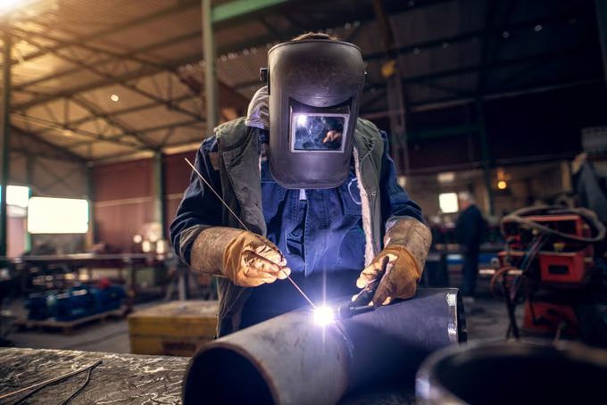 Welder in a flip-front helmet TIG welding a steel pipe elbow on a shop bench