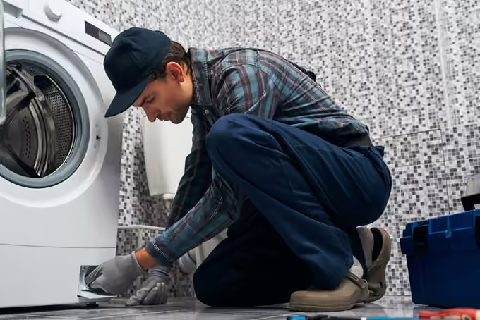 Appliance repair technician crouched at the filter door of a front-load washing machine with a toolbox