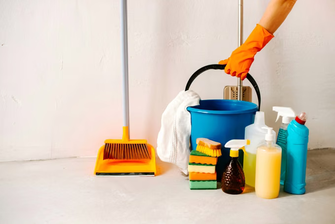 Cleaner's bucket, broom, sponges, and spray bottles lined up against a white wall