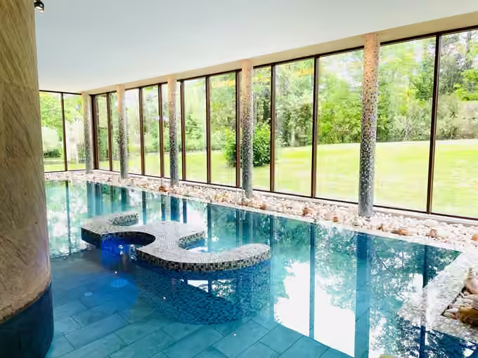 Indoor tiled pool with a built-in spa bench and floor-to-ceiling windows onto a lawn